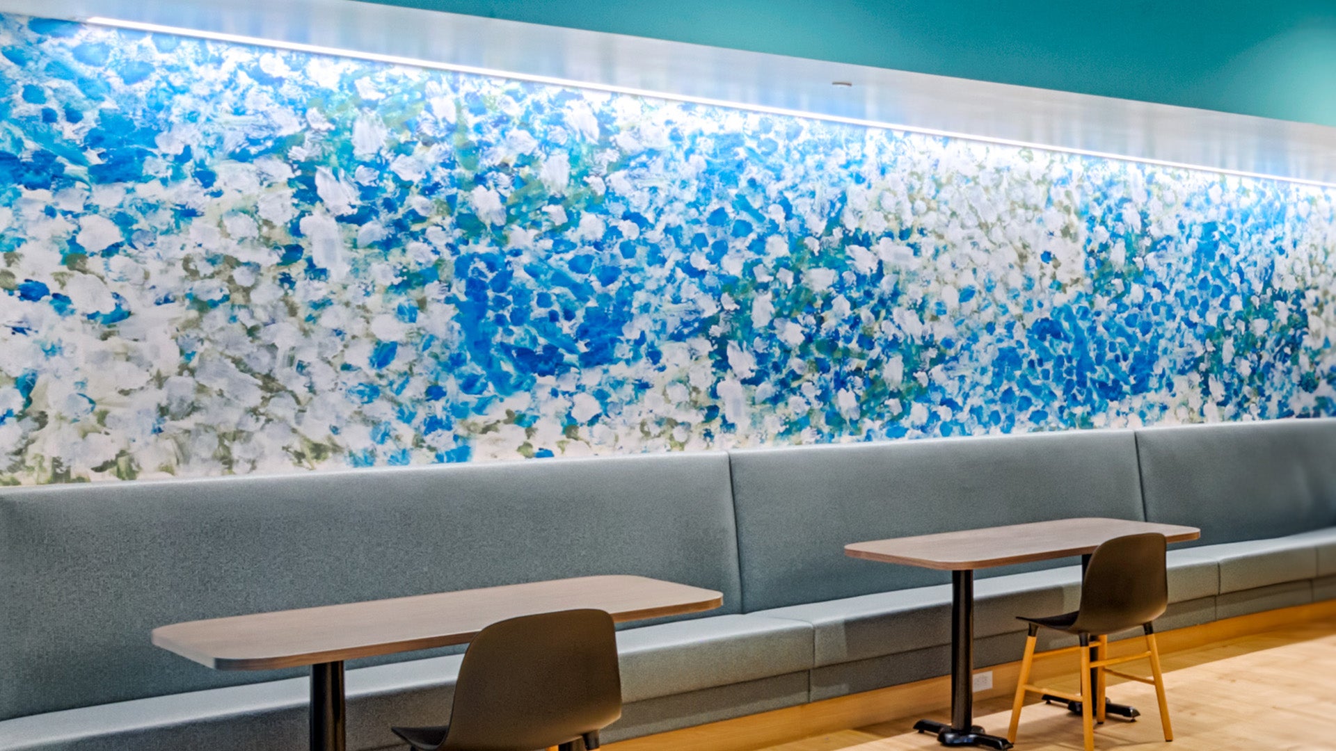 An abstract blue, white, and green painting by Mia Brown is reproduced as a wallcovering installed above a long bench in a cafe area in a tech office.