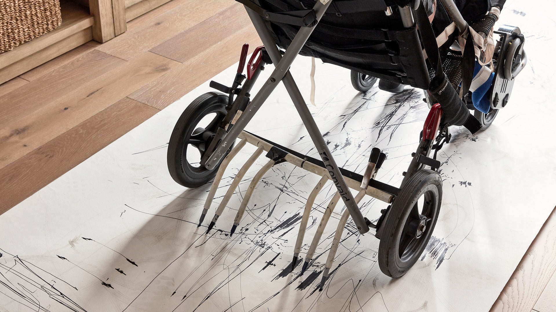ArtLifting artist Eric Santamaria from Los Angeles, is seated in his adaptive wheelchair with black markers attached to the back wheels. Eric is using his adaptive chair to make marks with black ink on a canvas laying on the floor. Photo courtesy Pottery Barn,