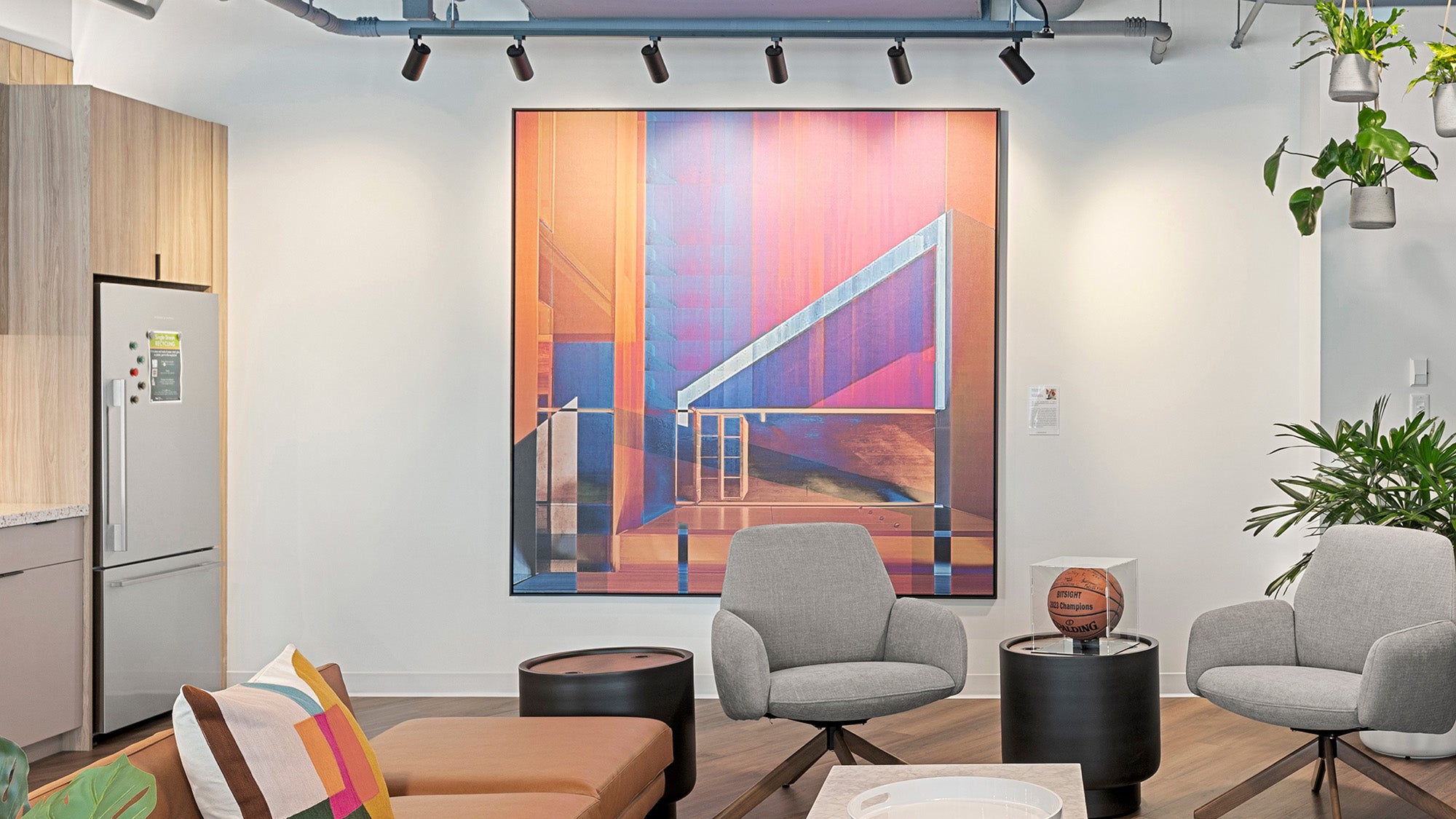 A huge artwork by Rick Ruark depicting a modern urban scene hangs on a white wall in an office common area with grey chairs, a leather couch, a stainless steel refridgerator, hanging plants, and a signed basketball displays on a side table.