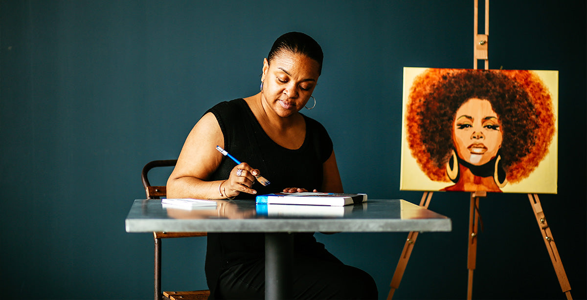 Artist Stacey Williams seated at a table painting on a canvas. A completed painting of Jill Scott is displayed on an easel next to her.