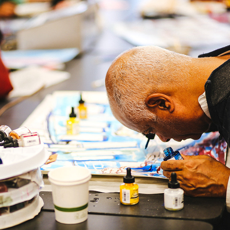 Artist Charles Blackwell leans close to a painting on a table and carefully applies ink with a dropper.