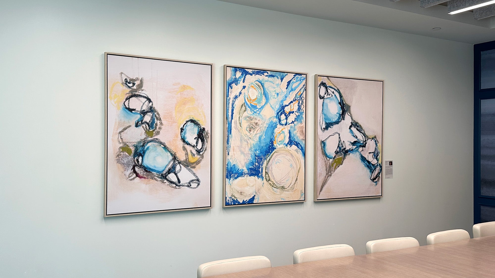 Three framed canvas prints by Elizabeth Gauss hang on a white wall in a conference room in a Zelis office in New Jersey. The triptych of artwork is abstract, looks sophisticated and abstract with blue, black, tan, and yellow elements. elements.