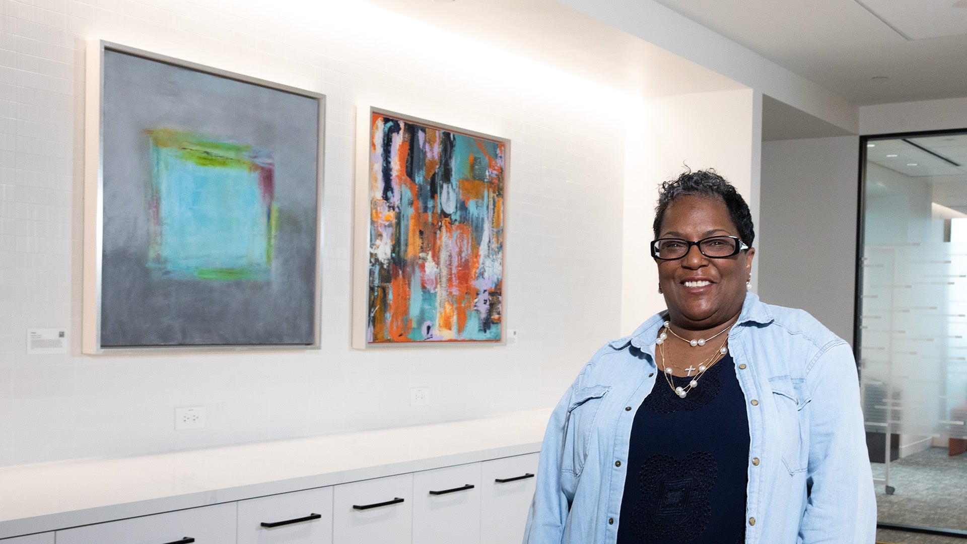 Artist Yvette stands smiling an office space for Wells Fargo, with her artwork behind her.